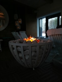 A decorative fire pit with glowing embers sits in a dimly lit room. It is surrounded by white stones arranged within a metal frame. In the background, there are chairs and a large circular mirror reflecting part of the room.