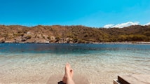 A landscape featuring a serene beach scene with a clear, turquoise shoreline in the foreground. A pair of bare feet is visible, suggesting relaxation or leisure. The background includes a range of rolling, forested hills under a bright, clear blue sky.