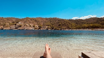 A landscape featuring a serene beach scene with a clear, turquoise shoreline in the foreground. A pair of bare feet is visible, suggesting relaxation or leisure. The background includes a range of rolling, forested hills under a bright, clear blue sky.