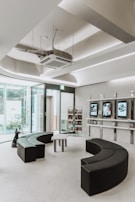 A modern, minimalist waiting room with semi-circular black seating arrangements and glass walls allowing natural light. There are display shelves with books and digital screens on the wall.