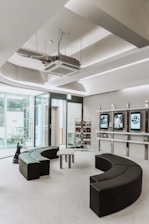 A modern, minimalist waiting room with semi-circular black seating arrangements and glass walls allowing natural light. There are display shelves with books and digital screens on the wall.
