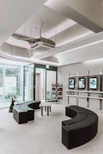 A modern, minimalist waiting room with semi-circular black seating arrangements and glass walls allowing natural light. There are display shelves with books and digital screens on the wall.