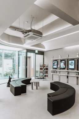 A modern, minimalist waiting room with semi-circular black seating arrangements and glass walls allowing natural light. There are display shelves with books and digital screens on the wall.