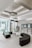 A modern, minimalist waiting room with semi-circular black seating arrangements and glass walls allowing natural light. There are display shelves with books and digital screens on the wall.