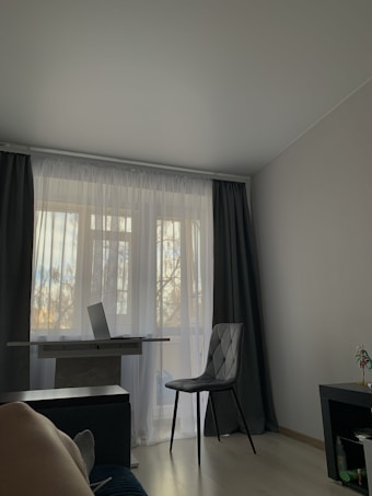 A minimalistic room with a window covered by white sheer curtains and dark drapes. In front of the window, there is a small desk with an open laptop on it. Next to the desk, there is a single dark-colored chair. The floor is light-colored, and there is a part of a leg resting on a piece of furniture in the foreground.