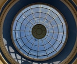 A circular glass dome featuring a grid of metal supports and a central design with concentric circles. The sky is visible through the clear glass panes, with light casting shadows through the structure onto the surrounding blue and beige framework.