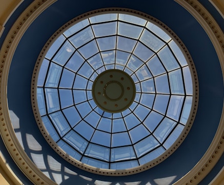 A circular glass dome featuring a grid of metal supports and a central design with concentric circles. The sky is visible through the clear glass panes, with light casting shadows through the structure onto the surrounding blue and beige framework.