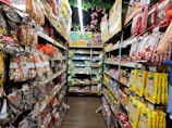A narrow grocery store aisle filled with colorful snack packages line both sides, including chocolate, chips, and candies. Bright packaging and vibrant signage are visible, with a ceiling adorned with decorative pink flowers.