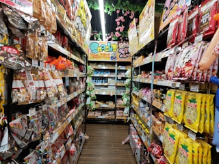 A narrow grocery store aisle filled with colorful snack packages line both sides, including chocolate, chips, and candies. Bright packaging and vibrant signage are visible, with a ceiling adorned with decorative pink flowers.