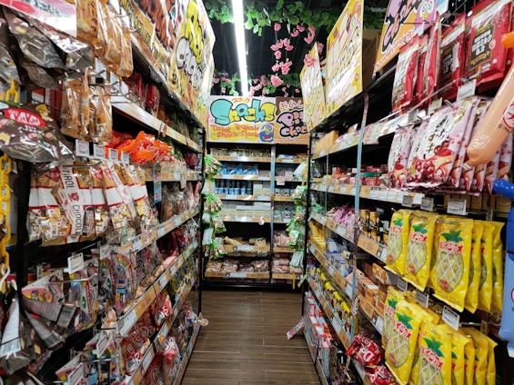 A narrow grocery store aisle filled with colorful snack packages line both sides, including chocolate, chips, and candies. Bright packaging and vibrant signage are visible, with a ceiling adorned with decorative pink flowers.