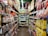 A narrow grocery store aisle filled with colorful snack packages line both sides, including chocolate, chips, and candies. Bright packaging and vibrant signage are visible, with a ceiling adorned with decorative pink flowers.