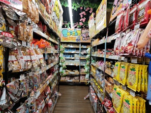 A narrow grocery store aisle filled with colorful snack packages line both sides, including chocolate, chips, and candies. Bright packaging and vibrant signage are visible, with a ceiling adorned with decorative pink flowers.