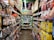 A narrow grocery store aisle filled with colorful snack packages line both sides, including chocolate, chips, and candies. Bright packaging and vibrant signage are visible, with a ceiling adorned with decorative pink flowers.