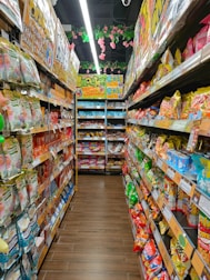 A colorful aisle in a store is lined with various packaged snacks on both sides. Brightly colored bags and boxes create a vibrant and appealing display. Overhead, pink flowers and green leaves hang decoratively from the ceiling. The shelves are stocked with different snack brands, featuring a wide range of colors and designs.