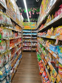 A colorful aisle in a store is lined with various packaged snacks on both sides. Brightly colored bags and boxes create a vibrant and appealing display. Overhead, pink flowers and green leaves hang decoratively from the ceiling. The shelves are stocked with different snack brands, featuring a wide range of colors and designs.