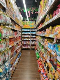 A colorful aisle in a store is lined with various packaged snacks on both sides. Brightly colored bags and boxes create a vibrant and appealing display. Overhead, pink flowers and green leaves hang decoratively from the ceiling. The shelves are stocked with different snack brands, featuring a wide range of colors and designs.