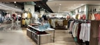 A well-lit clothing store interior featuring neatly arranged racks of clothing. There are various sections with diverse apparel including shirts, jackets, and pants in multiple colors. The center displays handbags and a table with folded clothes. The store has a modern design with wooden flooring and spotlights on the ceiling.