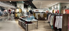 A well-lit clothing store interior featuring neatly arranged racks of clothing. There are various sections with diverse apparel including shirts, jackets, and pants in multiple colors. The center displays handbags and a table with folded clothes. The store has a modern design with wooden flooring and spotlights on the ceiling.