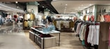 A well-lit clothing store interior featuring neatly arranged racks of clothing. There are various sections with diverse apparel including shirts, jackets, and pants in multiple colors. The center displays handbags and a table with folded clothes. The store has a modern design with wooden flooring and spotlights on the ceiling.