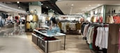 A well-lit clothing store interior featuring neatly arranged racks of clothing. There are various sections with diverse apparel including shirts, jackets, and pants in multiple colors. The center displays handbags and a table with folded clothes. The store has a modern design with wooden flooring and spotlights on the ceiling.