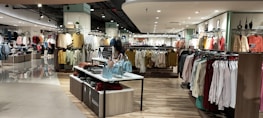 A well-lit clothing store interior featuring neatly arranged racks of clothing. There are various sections with diverse apparel including shirts, jackets, and pants in multiple colors. The center displays handbags and a table with folded clothes. The store has a modern design with wooden flooring and spotlights on the ceiling.