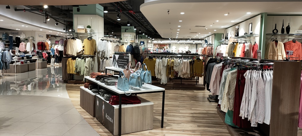 A well-lit clothing store interior featuring neatly arranged racks of clothing. There are various sections with diverse apparel including shirts, jackets, and pants in multiple colors. The center displays handbags and a table with folded clothes. The store has a modern design with wooden flooring and spotlights on the ceiling.