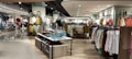 A well-lit clothing store interior featuring neatly arranged racks of clothing. There are various sections with diverse apparel including shirts, jackets, and pants in multiple colors. The center displays handbags and a table with folded clothes. The store has a modern design with wooden flooring and spotlights on the ceiling.