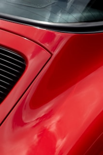 A close-up of a red car with glossy curved surfaces and a portion of the rear window. The vibrant red paint reflects clouds softly on the surface, and there is a black grille on the side.