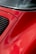 A close-up of a red car with glossy curved surfaces and a portion of the rear window. The vibrant red paint reflects clouds softly on the surface, and there is a black grille on the side.