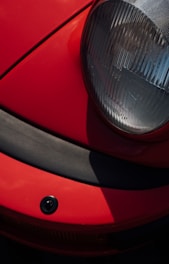 A close-up view of a red car's front end, featuring a prominently rounded headlight with a clear lens and a black rubber bumper below it. The image captures the car's sleek design with distinct shadows and highlights.