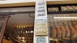 An Italian deli storefront featuring a glass window displaying hanging cured meats. Inside the shop, rows of sausages and hanging hams are visible alongside shelves lined with various bottles. Numerous signs with Italian text are displayed, listing products such as olive oil, pasta, and wine.