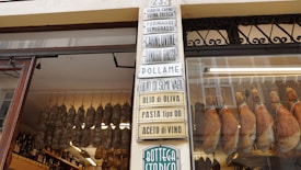 An Italian deli storefront featuring a glass window displaying hanging cured meats. Inside the shop, rows of sausages and hanging hams are visible alongside shelves lined with various bottles. Numerous signs with Italian text are displayed, listing products such as olive oil, pasta, and wine.