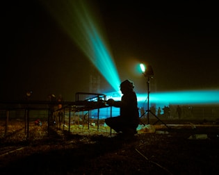 A silhouette of a person working with metal scaffolding under dramatic lighting. Bright beams of turquoise light cast across the scene, creating a striking contrast against the dark night sky. The environment suggests preparations for an outdoor event, with additional figures faintly visible in the background.