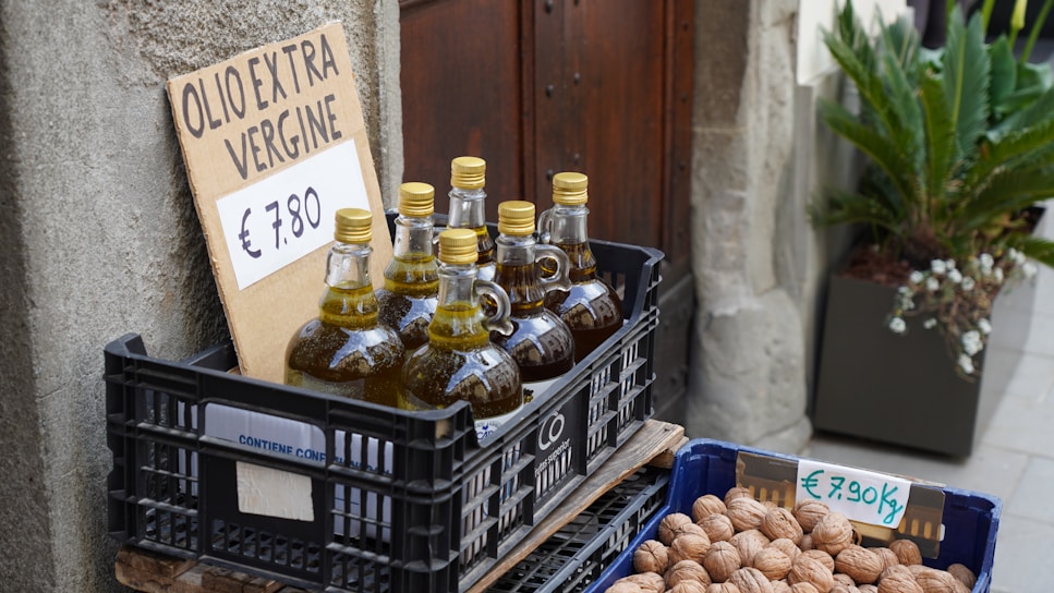Bottles of extra virgin olive oil are displayed in a plastic crate with a handwritten sign showing the price of €7.80. Next to it, there is another crate filled with walnuts, also with a price sign indicating €7.90 per kilogram. The setup is placed outdoors, possibly near a rustic wall with potted plants nearby.