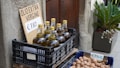 Bottles of extra virgin olive oil are displayed in a plastic crate with a handwritten sign showing the price of &euro;7.80. Next to it, there is another crate filled with walnuts, also with a price sign indicating &euro;7.90 per kilogram. The setup is placed outdoors, possibly near a rustic wall with potted plants nearby.