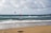 A sandy beach stretches into the distance, meeting the calm, blue ocean under a cloudy sky. Two kite surfers are visible in the water, with their colorful kites soaring above them. In the foreground, kiteboarding equipment is lying on the sand.