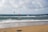 A sandy beach stretches into the distance, meeting the calm, blue ocean under a cloudy sky. Two kite surfers are visible in the water, with their colorful kites soaring above them. In the foreground, kiteboarding equipment is lying on the sand.