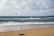 A sandy beach stretches into the distance, meeting the calm, blue ocean under a cloudy sky. Two kite surfers are visible in the water, with their colorful kites soaring above them. In the foreground, kiteboarding equipment is lying on the sand.