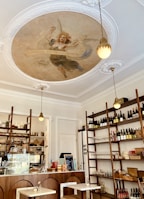 An elegant cafe interior with a vintage aesthetic. A large mural of a classical figure adorns the ceiling, surrounded by ornate white molding. Wooden shelves display an array of wine bottles, glasses, and other kitchen items. A small bar counter with a menu board and decorative items adds to the cozy atmosphere. Soft, warm lighting hangs from the ceiling, enhancing the inviting feel of the space.