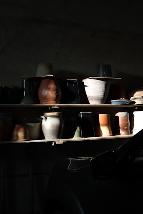 An assortment of ceramic pots and vases is displayed on a dimly lit wooden shelf. The objects vary in size and shape, with textures ranging from smooth to ribbed. Subtle light highlights certain pieces, creating shadows and an interplay of light and dark.