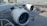 An airplane is parked at an airport with a focus on the two large jet engines prominently displaying a logo. The surrounding area includes tarmac, airport infrastructure, and a few small ground service vehicles.