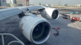 An airplane is parked at an airport with a focus on the two large jet engines prominently displaying a logo. The surrounding area includes tarmac, airport infrastructure, and a few small ground service vehicles.
