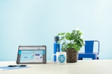 A light blue minimalistic setting featuring a tablet on a desk displaying a website, alongside a white mug with a blue logo, a woven pot with a leafy green plant, a blue reusable water bottle, and a blue metal chair in the background. A pen and a blue notebook are placed next to the tablet.