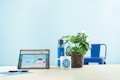 A light blue minimalistic setting featuring a tablet on a desk displaying a website, alongside a white mug with a blue logo, a woven pot with a leafy green plant, a blue reusable water bottle, and a blue metal chair in the background. A pen and a blue notebook are placed next to the tablet.