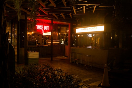 A night scene outside a food establishment that has two distinct signs: a glowing red 'TAKE OUT' sign with an arrow pointing downwards and a bright white sign that reads 'GREEN HOUSE'. The establishment has a somewhat rustic and cozy ambiance with subdued lighting inside. There are a few bar stools placed outside near a counter, and plants are partially visible in the foreground.