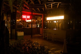 A night scene outside a food establishment that has two distinct signs: a glowing red 'TAKE OUT' sign with an arrow pointing downwards and a bright white sign that reads 'GREEN HOUSE'. The establishment has a somewhat rustic and cozy ambiance with subdued lighting inside. There are a few bar stools placed outside near a counter, and plants are partially visible in the foreground.