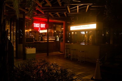 A night scene outside a food establishment that has two distinct signs: a glowing red 'TAKE OUT' sign with an arrow pointing downwards and a bright white sign that reads 'GREEN HOUSE'. The establishment has a somewhat rustic and cozy ambiance with subdued lighting inside. There are a few bar stools placed outside near a counter, and plants are partially visible in the foreground.
