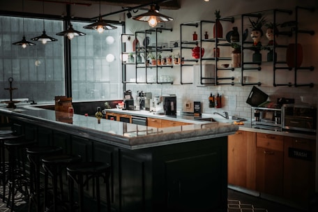 A cozy, dimly lit kitchen area featuring a long countertop with several bar stools. On the wall, there are shelves with various decorative items and plants. The kitchen is well-equipped with coffee machines, kitchen appliances, and utensils. Large windows allow natural light to filter through, giving the space a warm and inviting atmosphere.