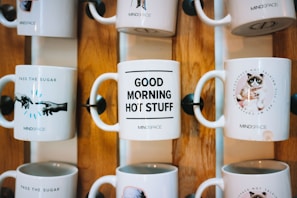 Several ceramic mugs are displayed on a wooden rack. Each mug features a different design and message. One mug reads 'PASS THE SUGAR' with an image of hands extended toward each other, another says 'GOOD MORNING HOT STUFF,' and a third features a grumpy cat with the message 'NOT TALKING TILL I GET MY COFFEE.' The mugs are arranged in two rows, showcasing a decorative and fun style.