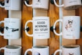 Several ceramic mugs are displayed on a wooden rack. Each mug features a different design and message. One mug reads 'PASS THE SUGAR' with an image of hands extended toward each other, another says 'GOOD MORNING HOT STUFF,' and a third features a grumpy cat with the message 'NOT TALKING TILL I GET MY COFFEE.' The mugs are arranged in two rows, showcasing a decorative and fun style.